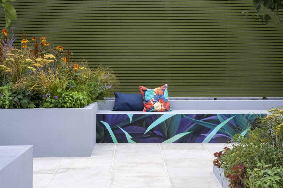 Grey bench with printed leaf design and raised beds with yellow-flowered plants edge Egyptian Beige Sawn Limestone paving slabs.