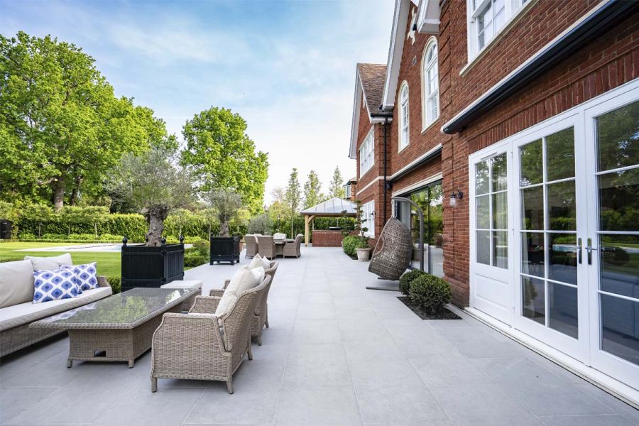 Florence Grey outdoor tiles UK stretch across width of house, with 2 sets of rattan furniture, olive tree planters and topiary.