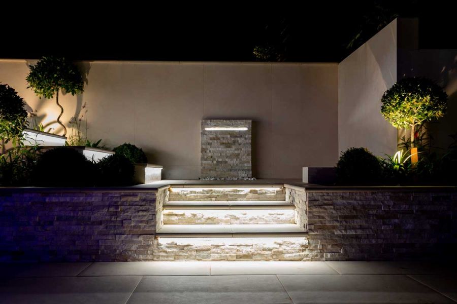 Underlit Venetian beige bullnose porcelain steps rise between stone-clad flanking walls to landing with illuminated monolith.