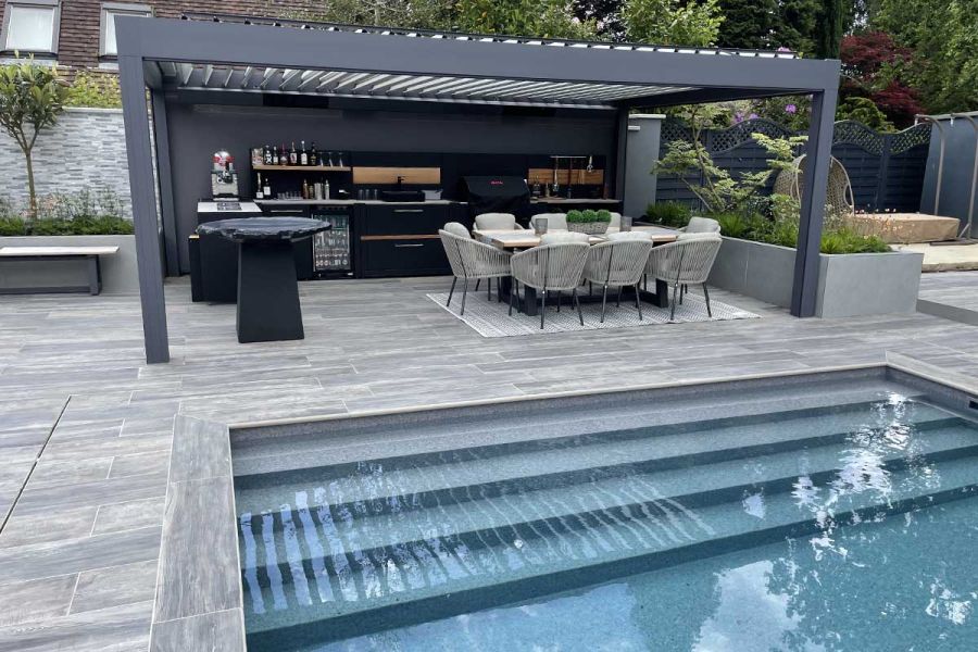 Beneath a spacious pergola, a cooking area with dining table. steps descend to the pool area, featuring Cinder Porcelain Paving.