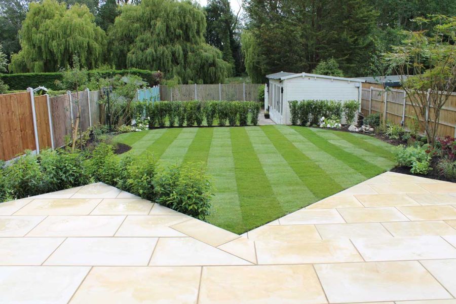 Geometrically edged patio of Harvest sawn sandstone slabs laid flush with striped lawn. Building behind hedge at end of garden.