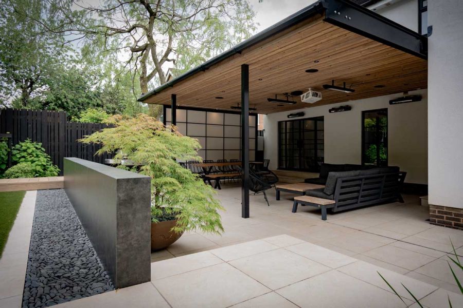 Covered paved patio divided from lawn by wall faced with dark grey Vulcano Roca external cladding in Japanese style garden.