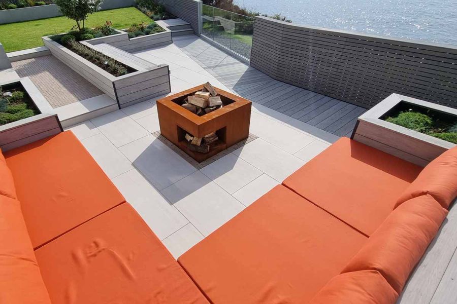 Faro porcelain patio with orange corner sofa and square fire pit next to rectilinear, multi-level raised beds. Lawn in background. 