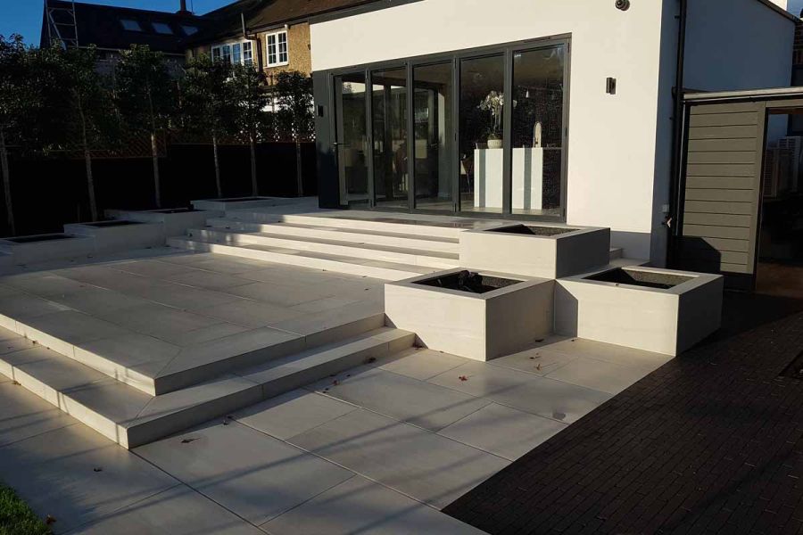 Steps descend from large glass french doors at back of house where Faro porcelain Paving is used as steps and paving surrounded by huge planters.