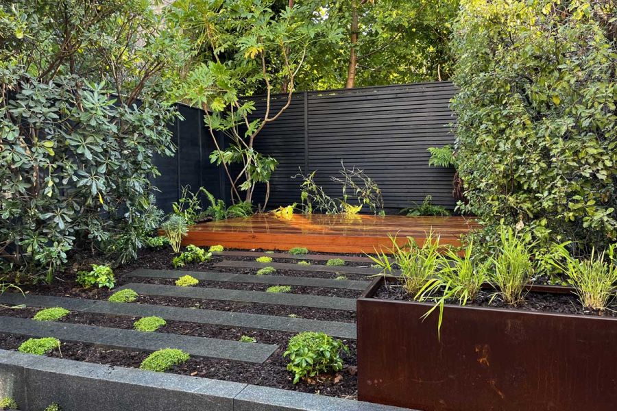 Raised area of Dark Grey granite plank paving alternating with strips of soil with low planting. Long wooden bench at back.