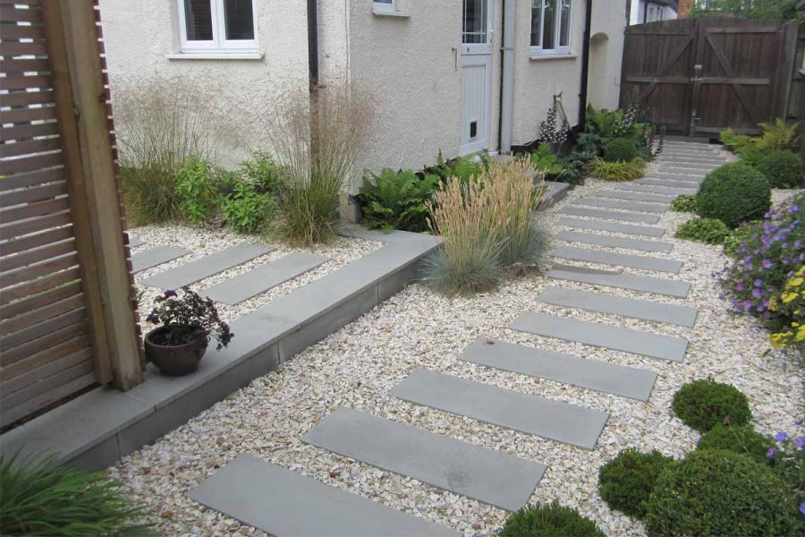 Flamed Grey Sawn Sandstone paving in short plank format, set in gravel, winds past ferns, grasses and box balls at back of house.