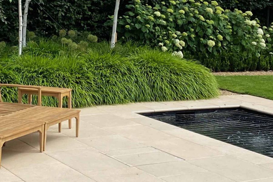 Florence beige porcelain paving with coping stones for garden walls used round edge of rectangular pond.  Grasses in background.
