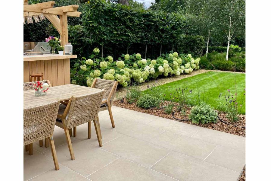Outdoor dining area using Florence Beige Porcelain Paving on the patio with well maintained lawn and plants opposite.