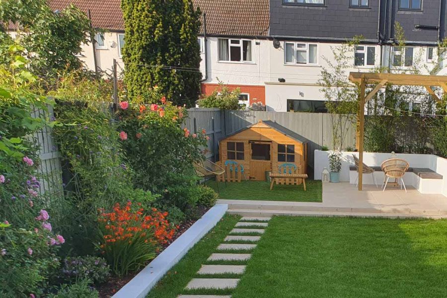 Path of Florence beige porcelain paving slabs leads down lawn to pergola-covered patio and Wendy house at end of fenced garden. 