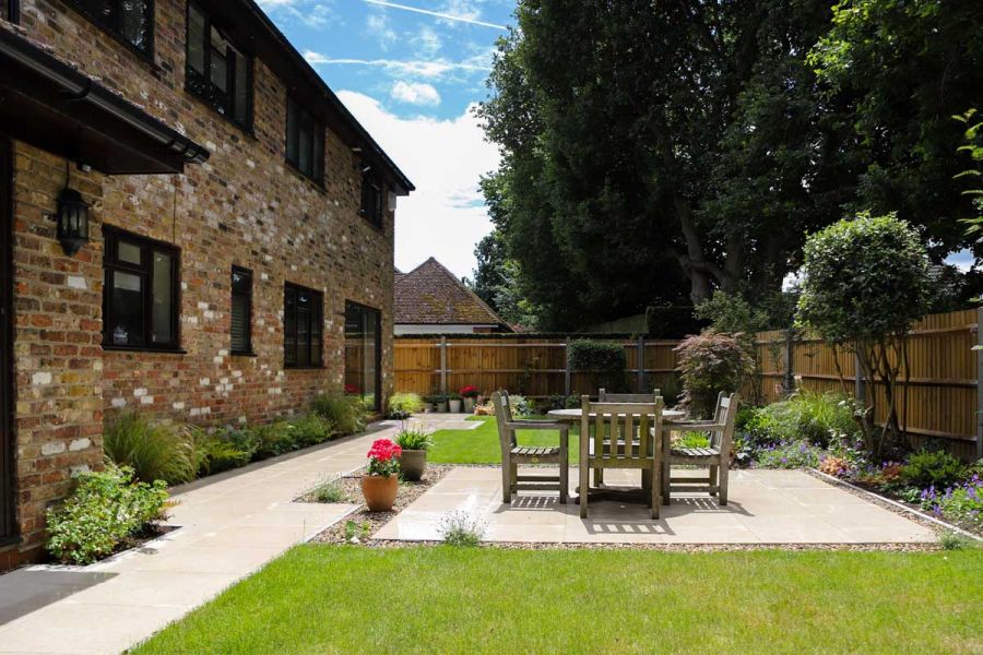 Wooden garden table and chairs on a Florence Beige Porcelain patio set in the middle of lawns and flower beds in a large rear garden.