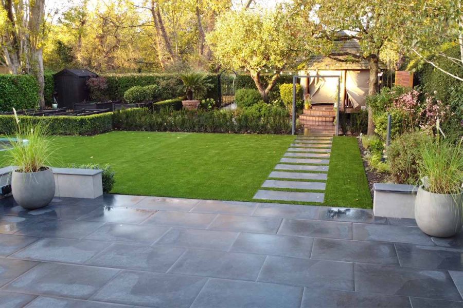 Florence Dark porcelain paving slabs cross lawn from matching patio to paved hedged area with raised beds. Design by Lucy Bravington.