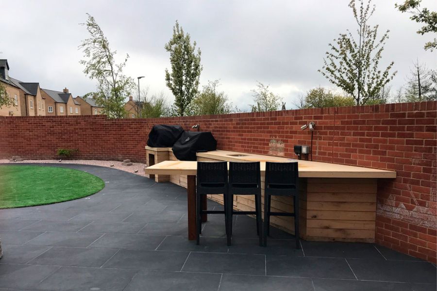 Outdoor kitchen against garden wall. Protruding bar with 3 stools. Florence Dark porcelain paving, by Muddy Boots Landscaping.
