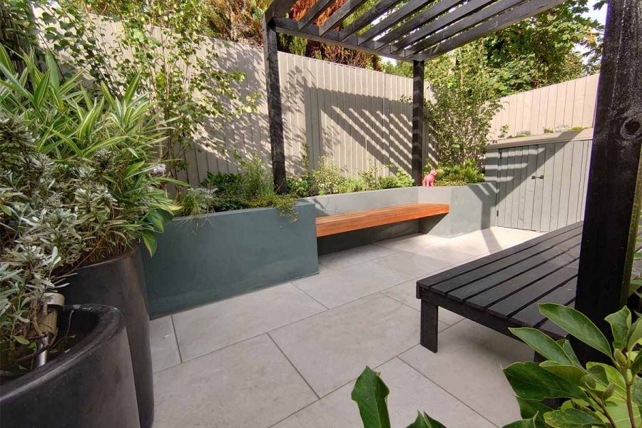 Small, tall-fenced outdoor space lined with raised beds and tall pots, tiled with Florence Grey porcelain under a wooden pergola.