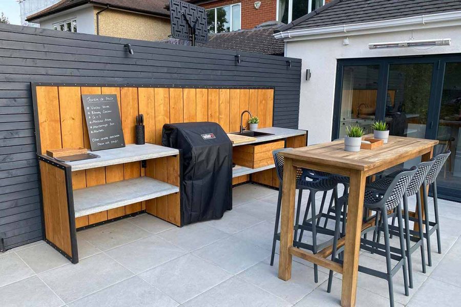 Patio of Florence Grey porcelain tiles, with bar table, 6 stools and outdoor kitchen. Drinks menu on blackboard on counter.