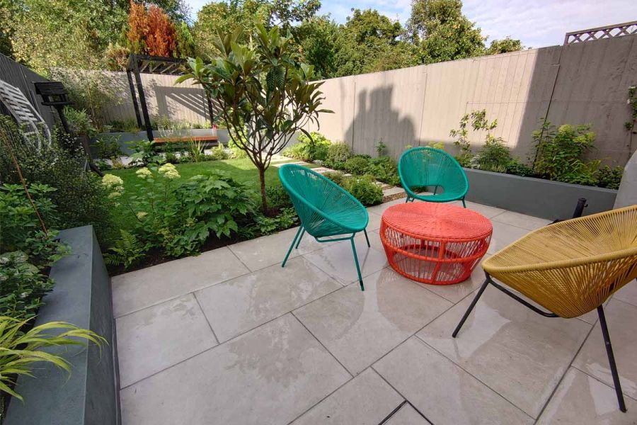 In fenced garden, colourful chairs and table sit on Florence Grey porcelain patio between grey-walled raised beds with lawn beyond.