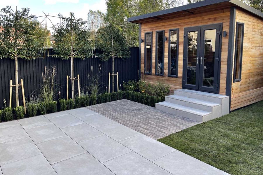 A wooden garden building with black trims sits at the end of florence grey porcelain paving patio, with steps leading up.