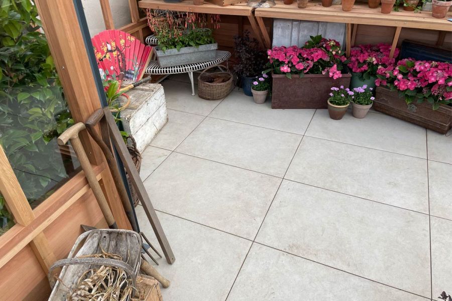 Florence Grey porcelain floor of wood-framed glasshouse, with gardening paraphernalia and troughs and pots of flowering plants.