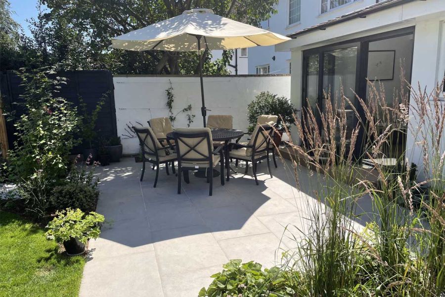 Sunny Florence Grey porcelain paved patio with 6-seat round dining set and parasol, edged by house, lawn, white wall and planting.