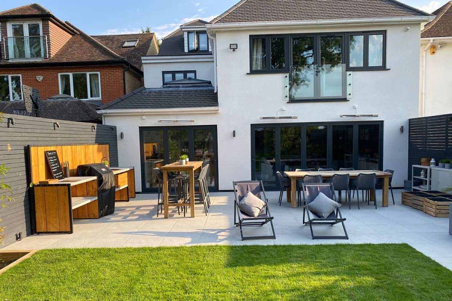 Sun lounges, bar set, outdoor kitchen and dining set sit on Florence grey porcelain paving patio at back of modern white house.