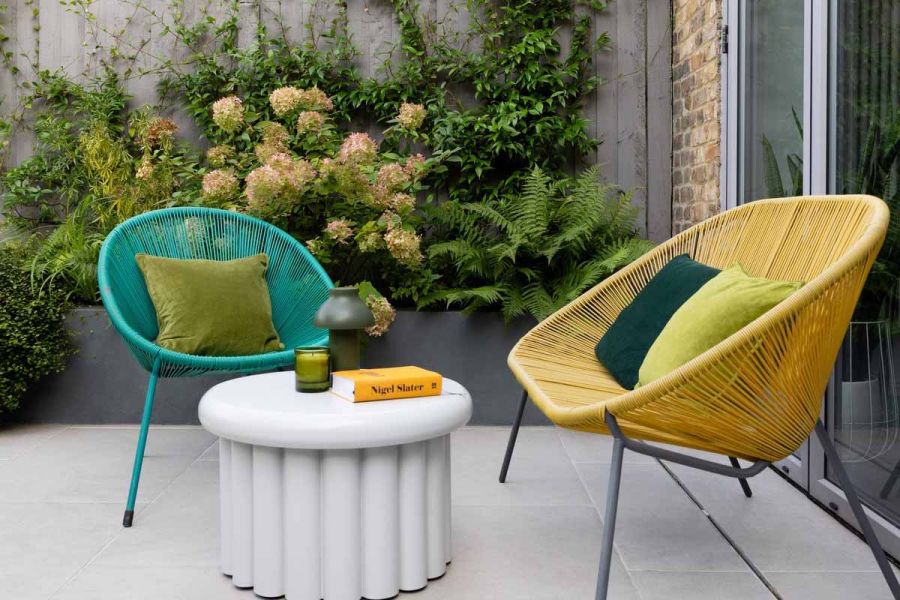 Florence Grey Porcelain Paving used as patio hosting unique shaped table with 2 colourful wire chairs and cushions sat against flowerbed.