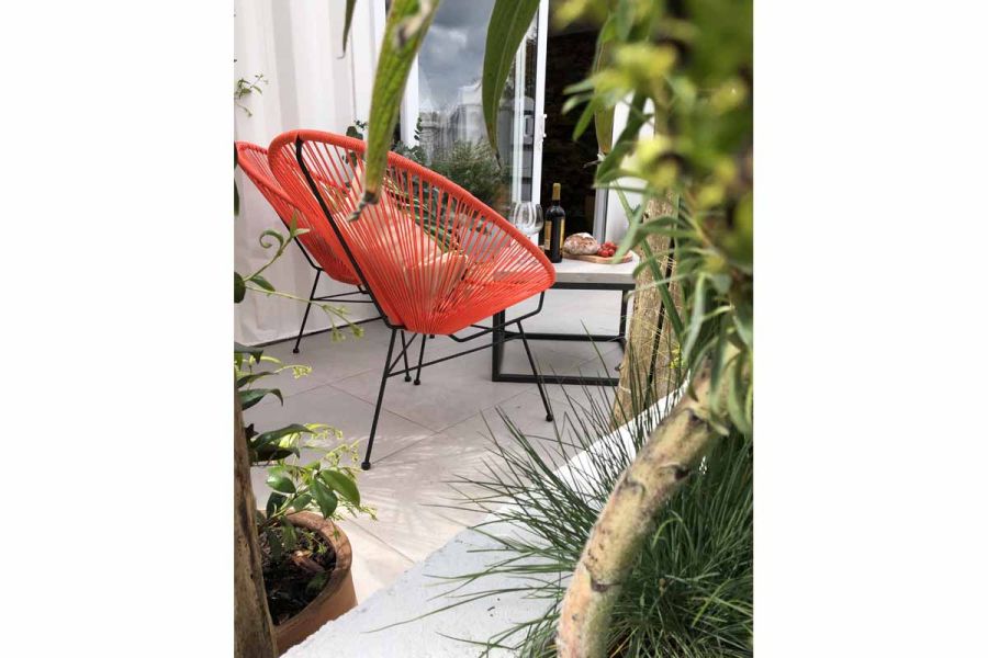 Orange and black wire chairs with coffee table on Florence Grey porcelain paving patio, seen through foliage, over low white wall.