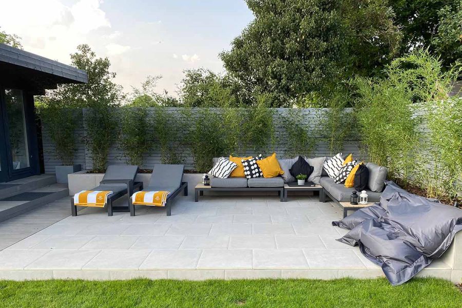 Raised patio of Florence Grey porcelain paving, with sunloungers, corner sofa and bean bags. Young trees line the edge on 2 sides.
