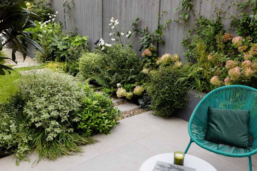 Lots of lush green planting surround a florence grey patio where a green wire chair sits along with pillow.
