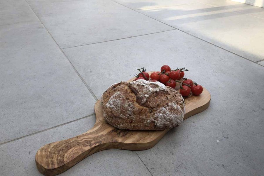 Paddle-shaped wooden board with loaf and tomatoes sits in shade on Florence Grey vitrified porcelain paving, laid in running bond.