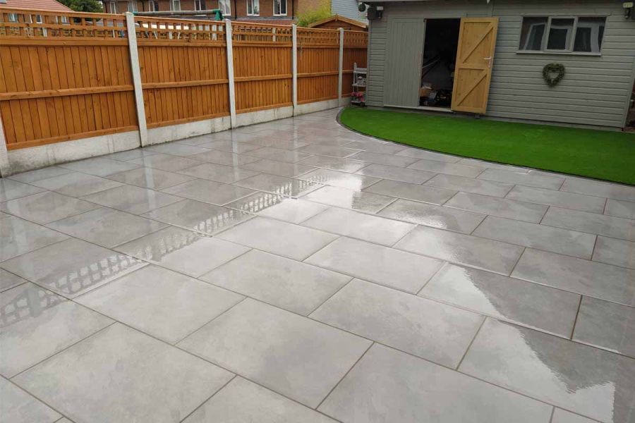 Wet Florence Storm porcelain paving laid running bond in fenced garden, edging small curved lawn in front of shed with door open.