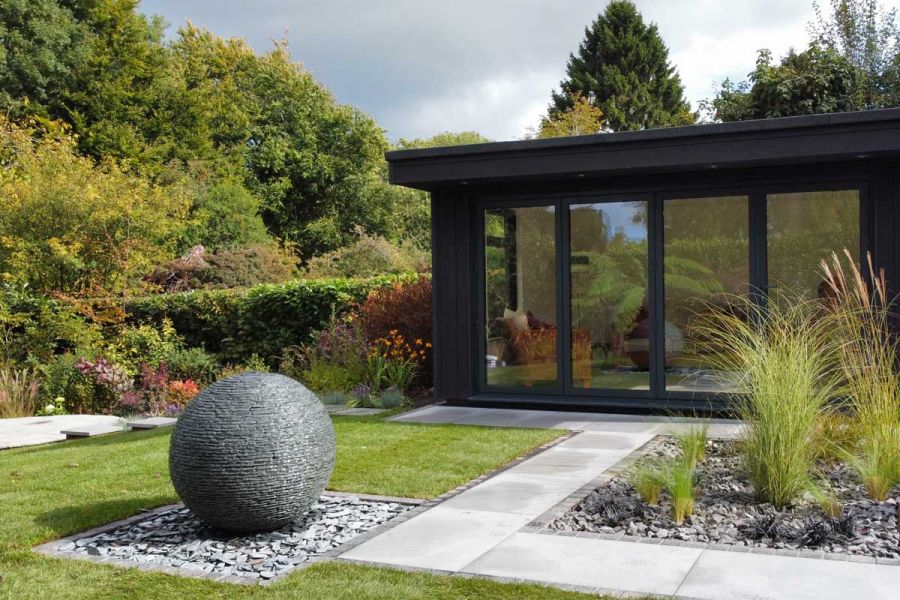 Mature rear garden featuring a modern glass fronted home office, grey porcelain paving paths and a large floating ball water feature.