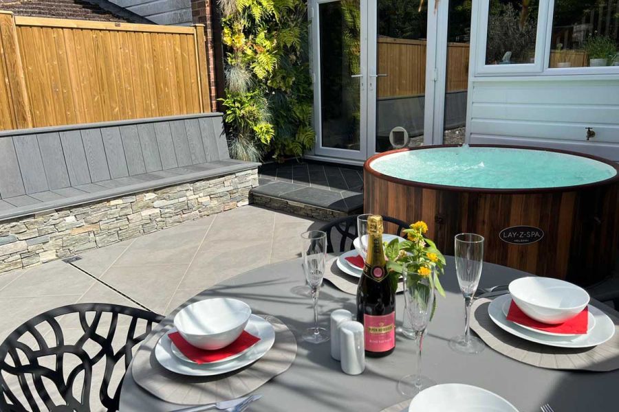 Table laid with crockery, glasses and bubbly in an intimate garden setting featuring a circular hot tub, built in bench and grey patio.