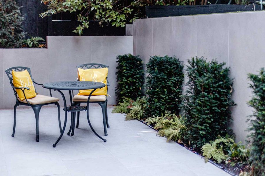 Metal bistro set with yellow cushions on Florence White porcelain paving, next to yew pillars. Design by Gardens of the Future.