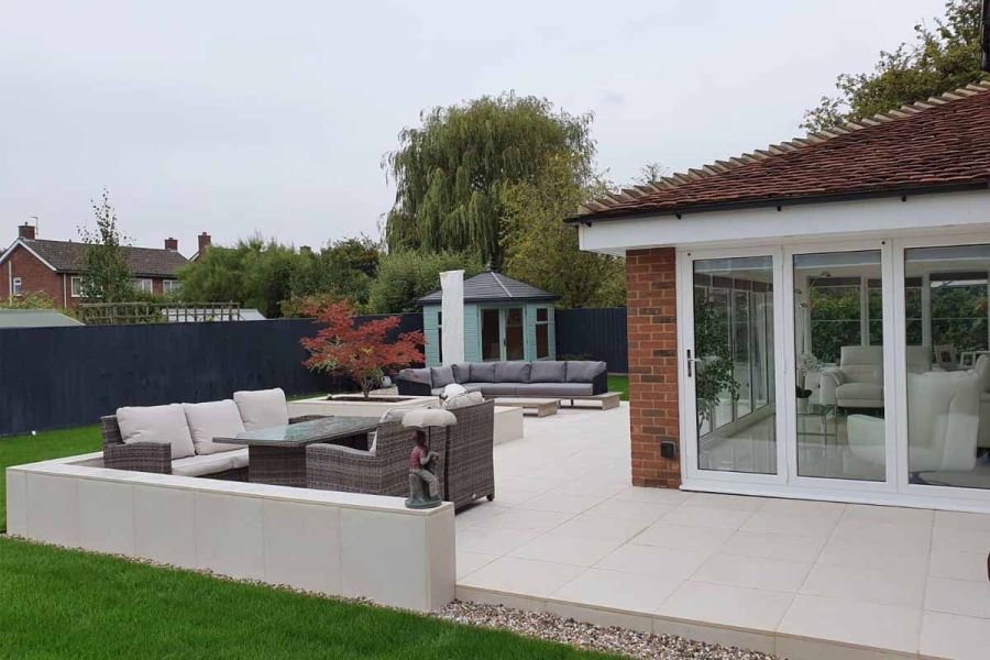 Low wall, faced with Florence White Porcelain slabs, divides lawn from matching patio wrapped around sunroom at back of house.