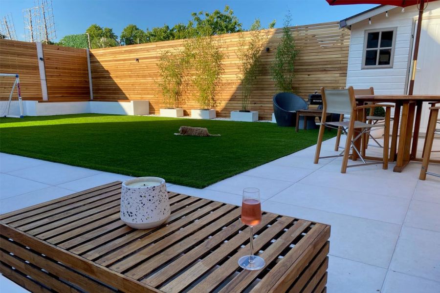 Dog lies on lawn by Florence White porcelain paving patio. Wine glass sits on slatted-wood coffee table next to garden furniture.