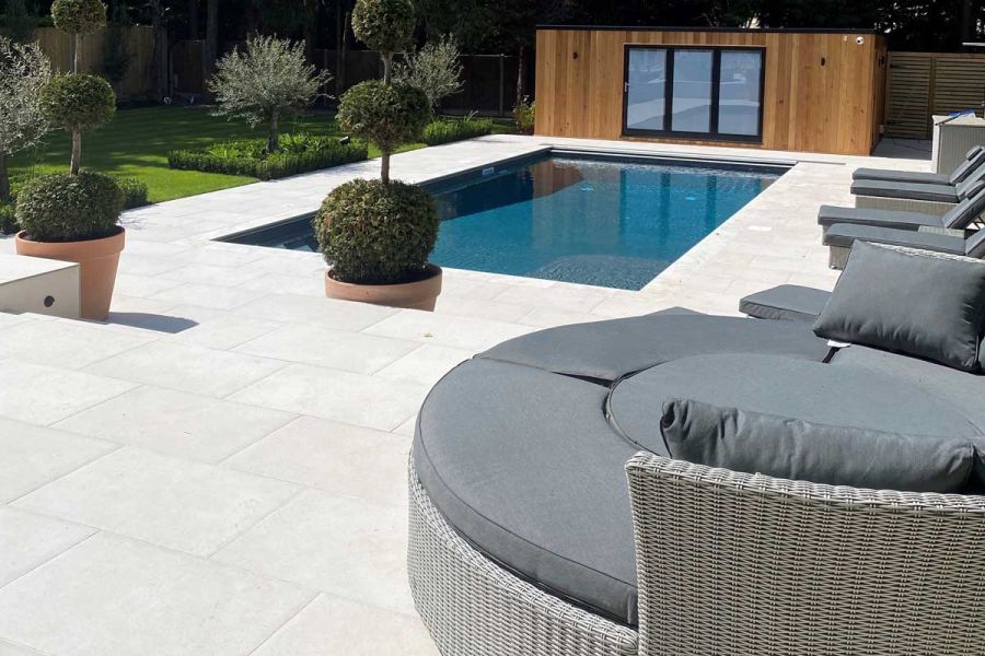 Florence white porcelain paving surrounds a swimming pool with planted trees and rattan furniture on the patio, garden building in the background.