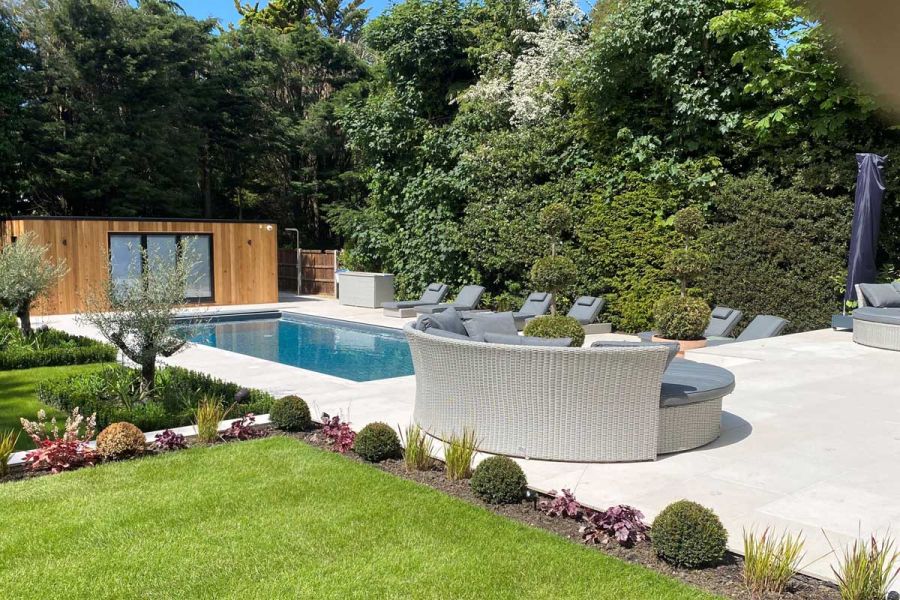 Florence White outdoor porcelain tiles UK surround swimming pool outside garden building. Round rattan couch on matching patio.
