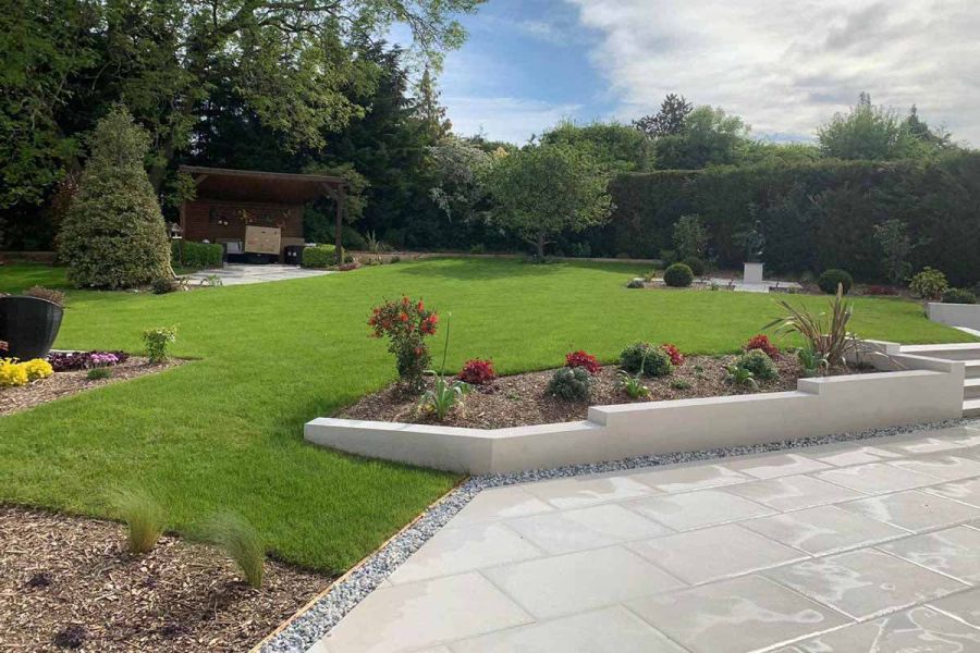 Low white wall retains flowerbed on edge of large lawn. Strip of grey pebbles at its base edges Florence White porcelain paving.