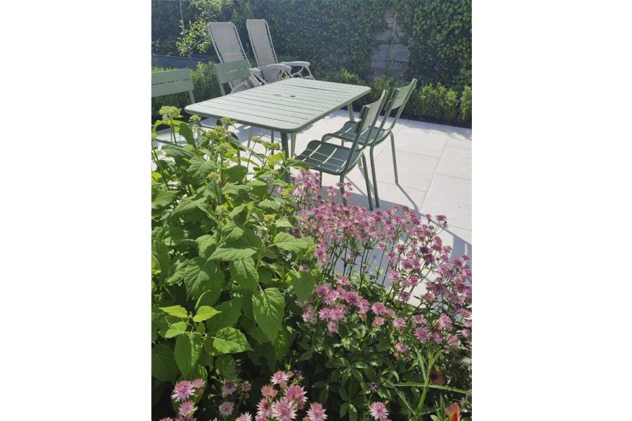View over bushy plants to green metal garden furniture and two recliners sitting on Florence white porcelain paving.