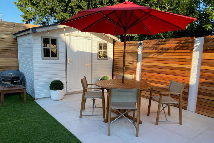 Wooden outdoor table, parasol and 4 folding chairs in front of white- painted shed, standing on Florence white porcelain paving.