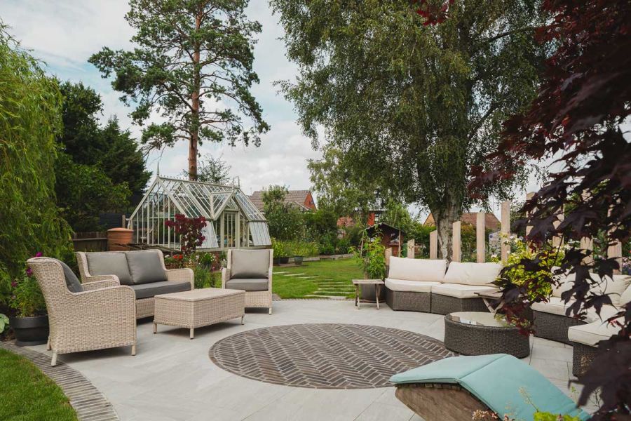 Moderna brick paving and Kandla Grey porcelain patio with outdoor furniture at bottom of garden with glasshouse nearby.
