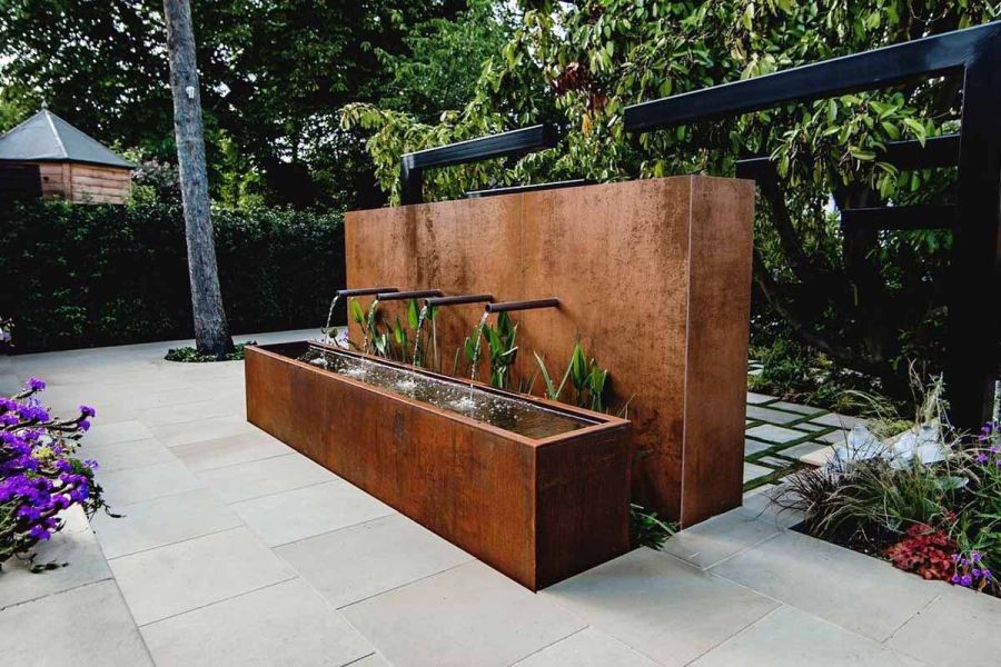 Corten steel water feature with water dribbling from 4 pipes into trough in garden corner paved with Britannia Buff sawn Yorkstone.