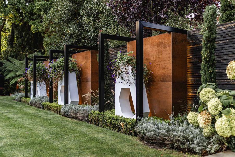 In planted border, 3 white sculptural planters set in front of tall blocks faced with Steel Corten external porcelain cladding.