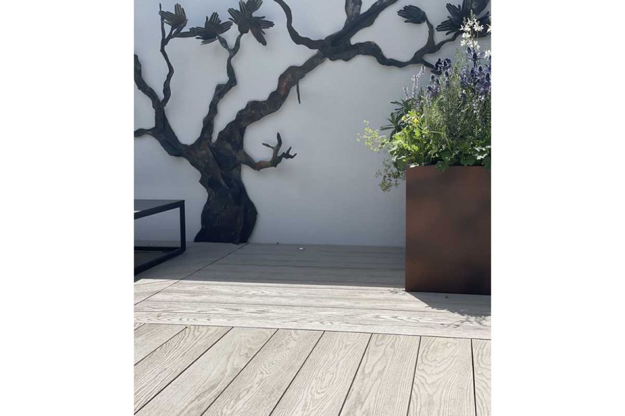 Corten steel planter sits on Limed Oak Millboard decking laid in 2 directions in front of white wall with metal silhouette of tree.