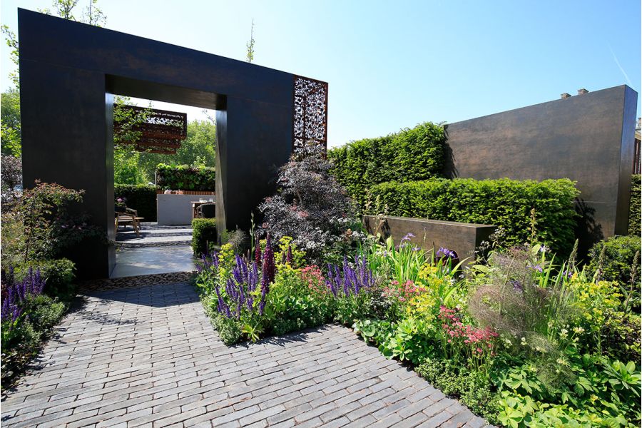 Square arch faced in Steel Dark porcelain cladding stands over wide path of clay pavers between deep planted beds.