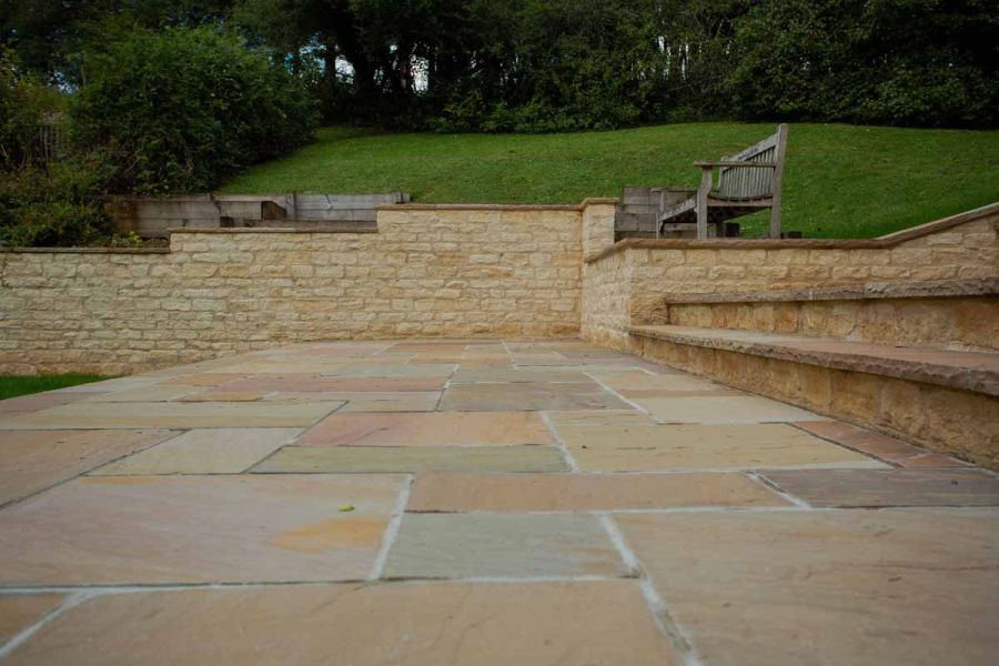 Sand-coloured retaining wall along far side of Camel Dust Indian sandstone patio, with 2 steps rising to right. 