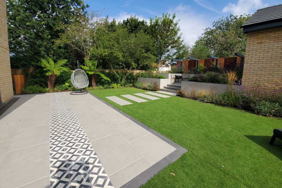 Path of Florence Grey Porcelain slabs crosses lawn from patio in matching paving and decorative tiles, to raised seating area.