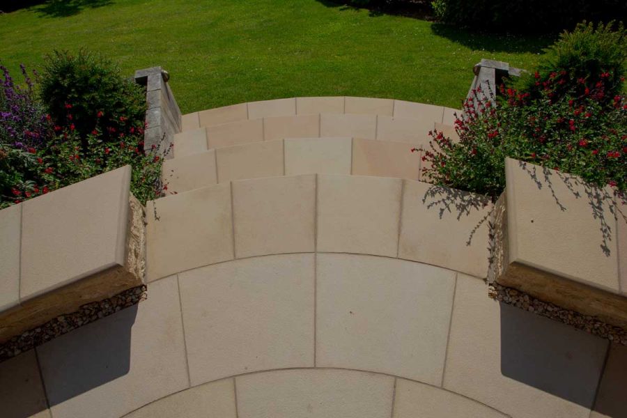 Bespoke curved buff sawn sandstone bullnose steps descend to lawn from patio with low walls. Design by Garden Solutions, Stroud. 
