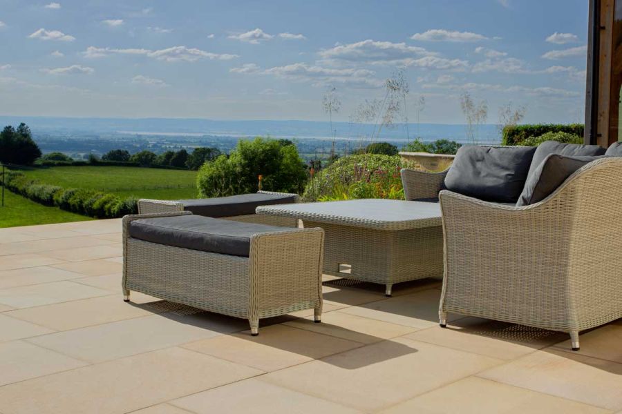 Rattan lounge set with dark grey cushions sits on edge of buff sawn paving terrace with view to blue horizon.