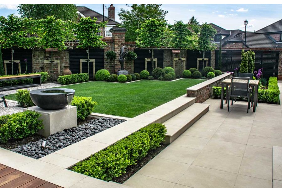 Garden by Robert Hughes, with Slab Khaki Chamfered Steps up from large matching patio to lawn with topiary and boxhead trees.