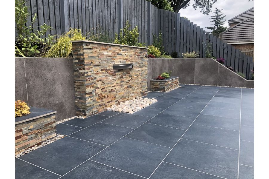 Charcoal porcelain paving patio in front of a stone clad water feature and large retaining wall clad with large format porcelain cladding.